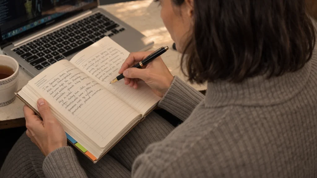 Close-up of hands writing a weekly study plan in a notebook beside a laptop
